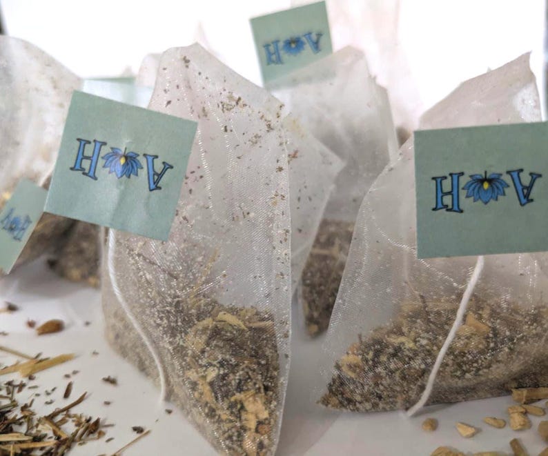May include: Several tea bags filled with loose leaf tea are arranged on a white surface. Each tea bag is made of a sheer, white material and has a small, light blue tag attached. The tags display the letters 'A' and 'H' for Astral Herbal with a blue lotus flower in between. The tea bags are surrounded by loose tea leaves and small pieces of ginger.