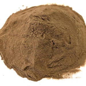 May include: A pile of fine, brown powder against a white background. The powder has a textured surface with subtle variations in color and tone. The image is well-lit, highlighting the details of the powder.