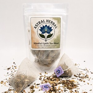 May include: A clear plastic bag of Astral Herbal Mindful Uplift Tea Blend with a white label featuring a blue lotus flower design. Two tea bags and loose tea leaves are scattered around the bag. The label lists ingredients like St. John's Wort and Ginger Root.