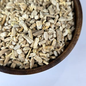 May include: Close-up of a wooden bowl filled with chopped, dried, light brown and beige sassafras root pieces. The pieces vary in size and shape, with some appearing like small sticks. The bowl has a dark brown rim and sits on a white surface.