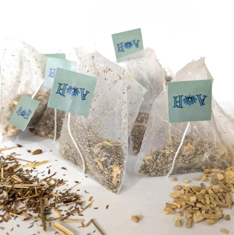 May include: Several tea bags filled with dried herbs and spices are arranged on a white surface. Each tea bag has a small, light blue tag with the letters "H" and "V" and a floral design. Loose herbs and spices are scattered around the tea bags.
