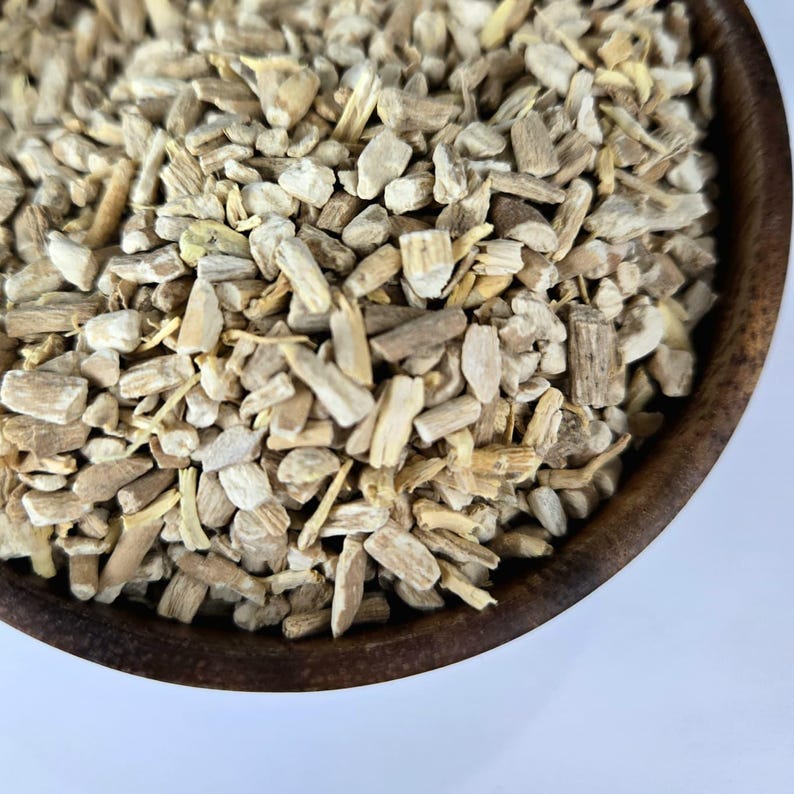 May include: Close-up of a wooden bowl filled with chopped, light brown and beige herbal pieces. The pieces have a rough, textured appearance, and vary in size and shape. The bowl is dark brown and sits on a white surface.