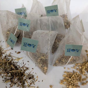 May include: A collection of tea bags filled with loose tea, each with a small tag featuring the letters "A H" and a floral design. Dried herbs and spices are scattered around the tea bags, suggesting a variety of tea blends.