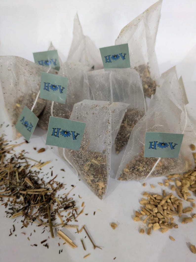 May include: A collection of tea bags filled with loose tea, each with a small tag featuring the letters "A H" and a floral design. Dried herbs and spices are scattered around the tea bags, suggesting a variety of tea blends.