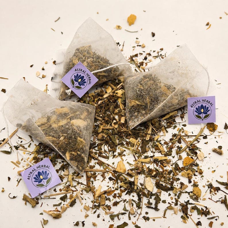 May include: Three tea bags filled with dried herbs and spices, with the text "ASTRAL HERBAL" on a purple tag. The tea bags are surrounded by loose herbs and spices. The tea bags are made of a translucent material.