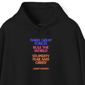 Puede incluir: Sudadera con capucha negra con bolsillo delantero y cordón. La parte delantera presenta la cita "THREE GREAT FORCES RULE THE WORLD STUPIDITY FEAR AND GREED" atribuida a Albert Einstein, en degradado azul, naranja y amarillo.