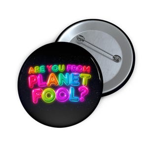 Op de afbeelding: Een zwarte button met de tekst "ARE YOU FROM PLANET FOOL?" in kleurrijke neonletters. De tekst is in rood, roze, geel, groen en blauw. De button heeft een glanzende metalen achterkant en een speld.