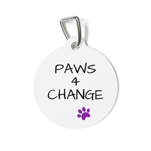 Puede incluir: Una etiqueta redonda blanca para mascotas con un lazo de metal plateado. La etiqueta presenta las palabras "PAWS 4 CHANGE" en negro, con una huella de pata morada debajo. La etiqueta está diseñada para mascotas.