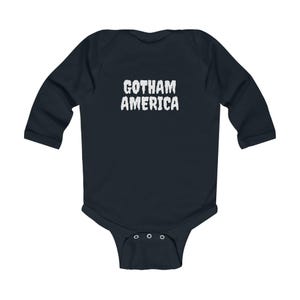 K&ouml;nnte beinhalten: Ein marineblaues Baby-Body mit langen &Auml;rmeln und dem wei&szlig;en Text "GOTHAM AMERICA" auf der Brust. Der Body hat einen Druckknopfverschluss am unteren Ende und ist aus weichem Material gefertigt.
