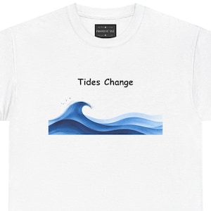 May include: A white short-sleeve t-shirt featuring a graphic of blue ocean waves and the text "Tides Change" printed in black above the waves. The shirt has a black inner neck label with the text "PROTEST TEE" in white lettering.