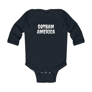 K&ouml;nnte beinhalten: Ein marineblaues Baby-Body mit langen &Auml;rmeln und dem wei&szlig;en Text "GOTHAM AMERICA". Der Body hat einen Druckknopfverschluss am unteren Ende und ist aus weichem Material gefertigt.