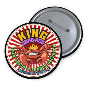 May include: A round button with a cartoon crab wearing a crown. The button has the text "KING GRAB ASS" and "WHEN YOU'RE A FAMOUS PREDATOR THEY LET YOU DO IT". The button is red, white, and black.