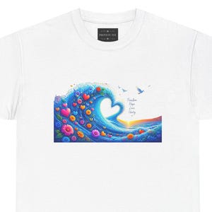 May include: A white t-shirt featuring a graphic of a blue ocean wave forming a heart shape, decorated with colorful flowers and hearts. The text reads Freedom, Hope, Love, Unity, with small birds flying above the water and a sunset on the horizon.