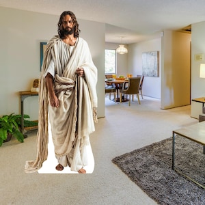 May include: A life-size cardboard cutout of a man with long brown hair and a beard, wearing a white robe and holding a draped cloth. The cutout is standing in a living room setting with a table, chairs, and a rug.