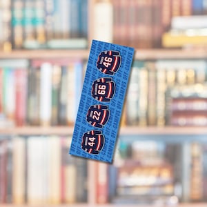 Op de afbeelding: Een blauwe boekenlegger met vier hockeytrui-ontwerpen met de nummers 44, 22, 66 en 46. De tekst "OFF-CAMPUS" is verticaal afgedrukt. De achtergrond is een vage boekenkast.