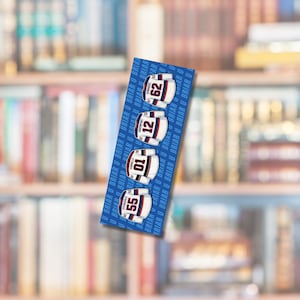 Op de afbeelding: Een blauwe boekenlegger met vier hockeytrui-ontwerpen met de nummers 55, 01, 12 en 62. De truien zijn wit met rode en blauwe accenten. De achtergrond is een wazige afbeelding van een boekenplank.