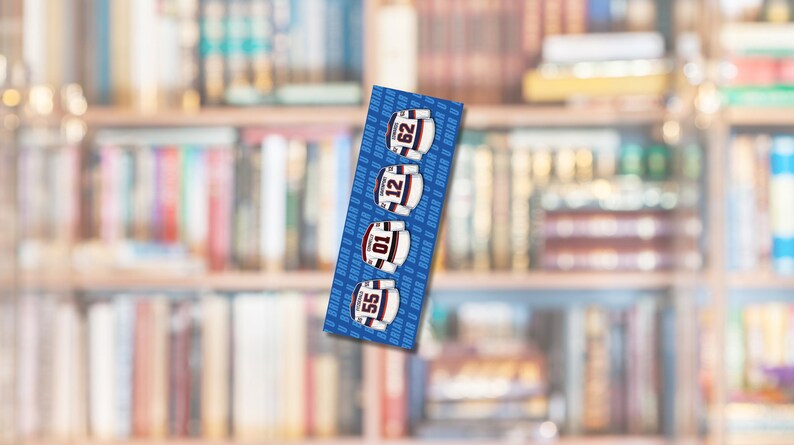 Op de afbeelding: Een blauwe boekenlegger met vier hockeytrui-ontwerpen met de nummers 55, 01, 12 en 62. De truien zijn wit met rode en blauwe accenten. De achtergrond is een wazige afbeelding van een boekenplank.