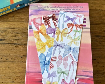 May include: Two decorative bookmarks with watercolor bow designs rest on a book. The bookmarks feature bows in various colors, including yellow, blue, and pink. The book has a pink cover with the title "Fortune" and author information.