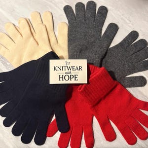 May include: A collection of knitted gloves in cream, dark grey, navy blue, and red arranged in a fan pattern. A rectangular white card is placed in the centre featuring the text KNITWEAR with HOPE and a small bird icon above the word KNITWEAR.