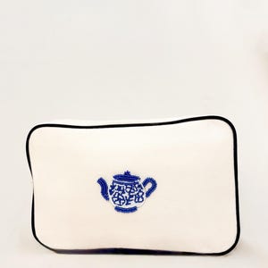 May include: A rectangular white pouch with a black trim. The pouch features a blue teapot design made of small beads. The teapot design is centered on the pouch.