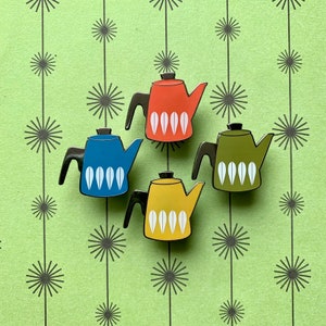 May include: Four enamel pin badges of vintage style coffee pots in red, blue, green and yellow with white accents. Each pot has a stylized design of white leaves on the side.