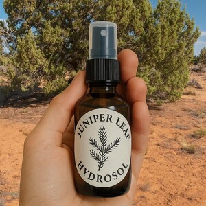 May include: A brown glass spray bottle labeled "Juniper Leaf Hydrosol" with a black cap, held against a backdrop of green trees and a blue sky. The label features a sprig of juniper. The bottle is held in a hand.