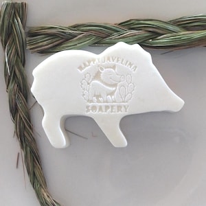 May include: A white soap shaped like a javelina, with the text "Happy Javelina Soapery" embossed on its surface. The soap is surrounded by braided and loose strands of dried grass. The image is set on a white plate.