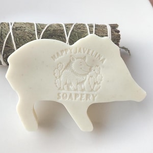 May include: A white, pig-shaped soap bar with the text "HAPPY JAVELINA SOAPERY" and a graphic of a javelina. A bundle of dried sage is tied with white string and rests behind the soap.