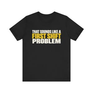 Könnte beinhalten: Ein schwarzes T-Shirt mit kurzen Ärmeln und dem Text "THAT SOUNDS LIKE A FIRST SHIFT PROBLEM" in einer abgenutzten Schriftart. Die Wörter "THAT SOUNDS LIKE A" und "PROBLEM" sind weiß, während "FIRST SHIFT" in einem kräftigen Gelb gehalten ist.
