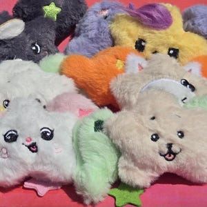 May include: A collection of plush star-shaped toys in various colors, including white, orange, yellow, and purple. Each star has embroidered facial features and a small star accent. The toys are soft and fluffy, with a playful and whimsical design.