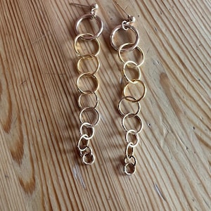 May include: A pair of gold-toned earrings. Each earring features a series of connected rings, creating a cascading effect. The earrings are displayed against a wooden background, highlighting their design and color.
