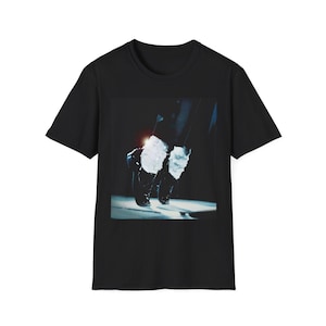 May include: Black t-shirt featuring a graphic of black shoes and white socks, with a stage light effect. The image is a close-up of the feet of a person doing a dance move. The shirt is a crew neck.