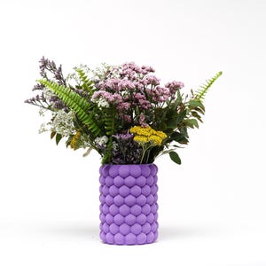 May include: A vibrant bouquet of flowers in a textured, lavender-colored vase. The arrangement includes a variety of blooms in shades of purple, yellow, and white, with green foliage. The vase has a unique, bubble-like design.