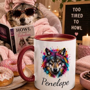 Peut inclure: Un mug en céramique blanche avec un intérieur et une anse rouge foncé, orné d'une illustration de tête de loup aux éclaboussures de peinture colorées. Sous le motif du loup, le prénom Penelope est inscrit en écriture cursive noire élégante.