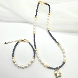 May include: A necklace and bracelet set featuring a combination of white pearls, black beads, and gold-colored accents. The necklace has a gold heart pendant. The jewelry is displayed on a white background.