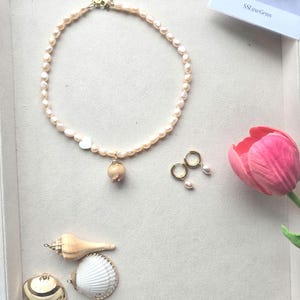 Puede incluir: Una colección de joyas y objetos decorativos, que incluye un collar de perlas, pendientes de aro dorados con perlas y conchas marinas. Un tulipán rosa está en la esquina. El collar tiene un dije en forma de corazón y un colgante.