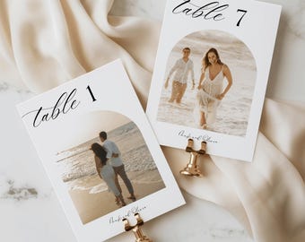 Puede incluir: Dos tarjetas de mesa blancas con fotos de una pareja en la playa, sujetas con clips dorados. Las tarjetas tienen las palabras "Table 1" y "Table 7" en escritura elegante. La parte inferior de las tarjetas tiene las palabras "Frank and Oliver".