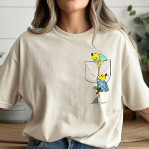 May include: Beige t-shirt with a pocket featuring a cartoon illustration of two bananas fishing. One banana is in the pocket, and the other is climbing down a rope towards a shark in the water. The bananas are wearing blue and yellow outfits.