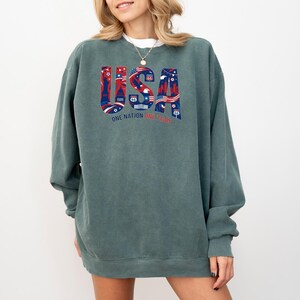 May include: A teal-colored sweatshirt with the word "USA" in a patriotic design. The letters are filled with red, white, and blue illustrations of American landmarks and symbols. The text "ONE NATION ONE TEAM" is below the word "USA".