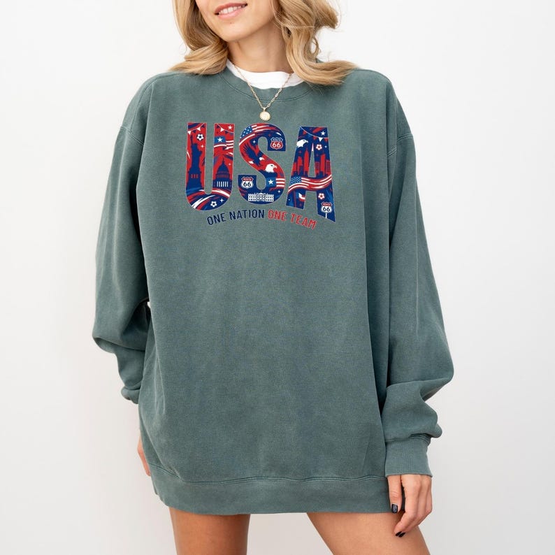 May include: A teal-colored sweatshirt with the word "USA" in a patriotic design. The letters are filled with red, white, and blue illustrations of American landmarks and symbols. The text "ONE NATION ONE TEAM" is below the word "USA".