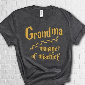 May include: Dark gray t-shirt with the words "Grandma manager of mischief" in gold lettering. The text is styled in a whimsical font with a trail of footprints. The shirt is made of a soft material.
