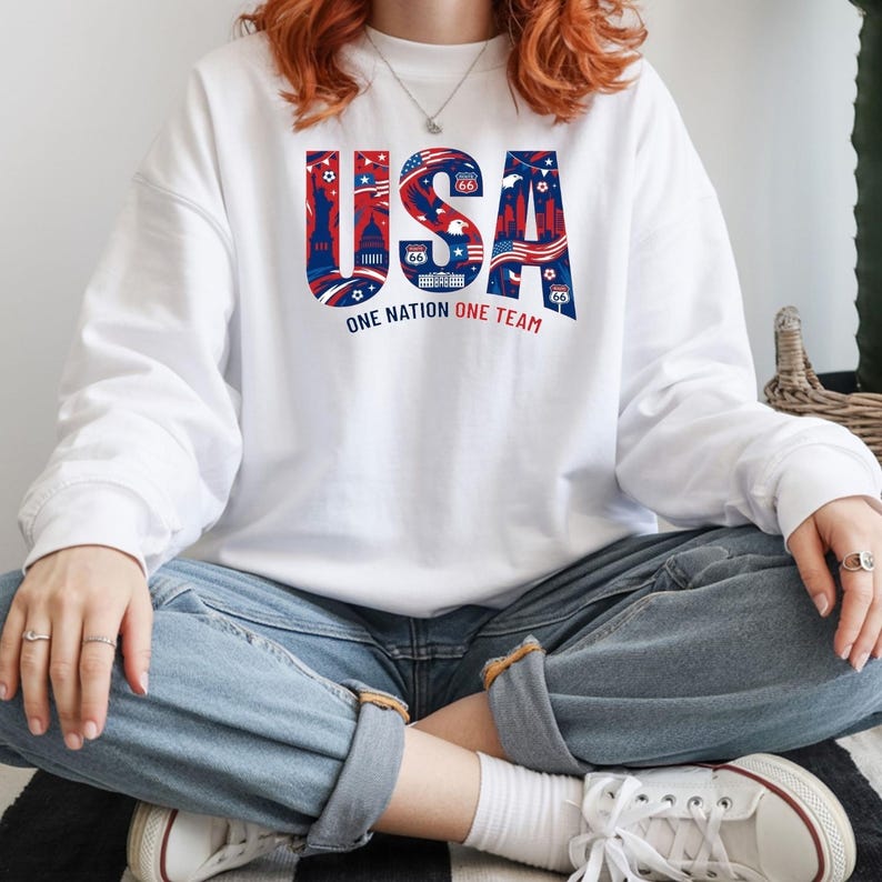 May include: White long-sleeve sweatshirt with a patriotic "USA" graphic in red, white, and blue. The graphic includes Route 66 imagery and the text "One Nation One Team". The sweatshirt is a casual style.