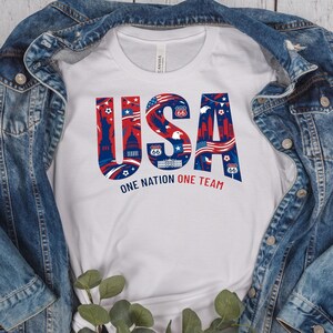 May include: White t-shirt with a patriotic design featuring the word "USA" in red and blue, filled with iconic American imagery. The text "ONE NATION ONE TEAM" is printed below the word "USA".
