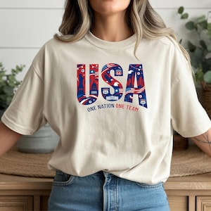 May include: A beige t-shirt with the word "USA" in red, white, and blue, featuring iconic American imagery. The text "ONE NATION ONE TEAM" is below the word "USA". The shirt has short sleeves and a crew neck.