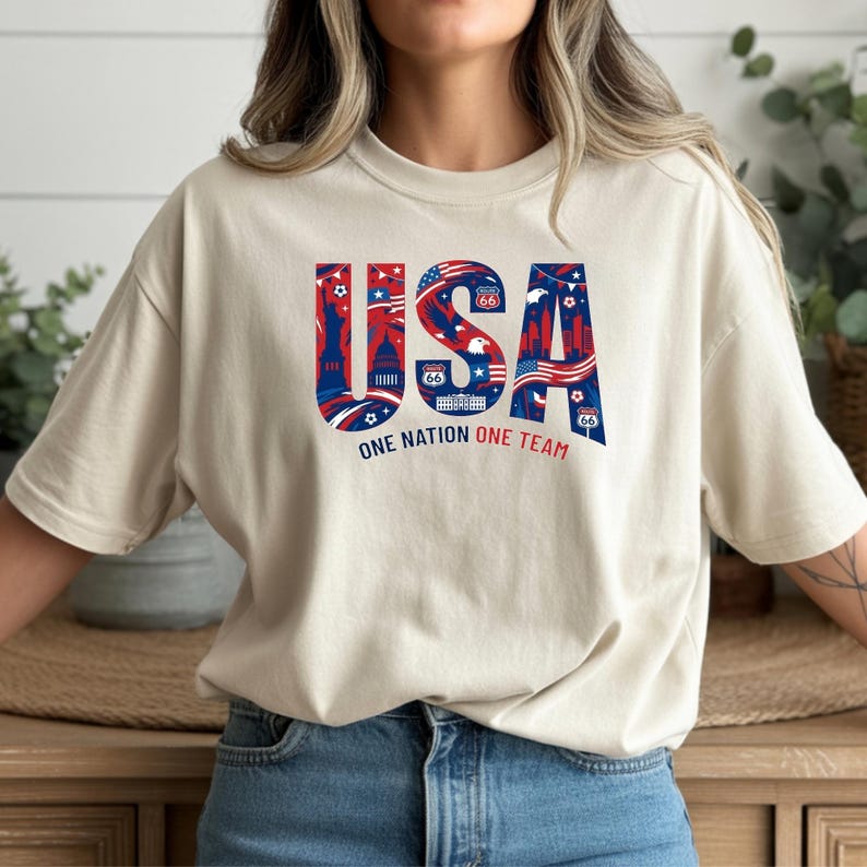 May include: A beige t-shirt with the word "USA" in red, white, and blue, featuring iconic American imagery. The text "ONE NATION ONE TEAM" is below the word "USA". The shirt has short sleeves and a crew neck.