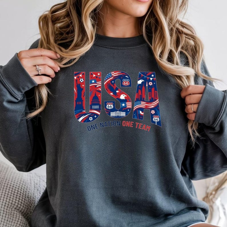 May include: Dark gray sweatshirt with a colorful "USA" graphic. The letters are filled with patriotic imagery, including the American flag, landmarks, and Route 66 signs. The text "ONE NATION ONE TEAM" is below the graphic.