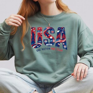 May include: A sage green sweatshirt with the word "USA" in red, white, and blue, featuring iconic American imagery. The text "ONE NATION ONE TEAM" is below the word "USA". The sweatshirt is a casual style.