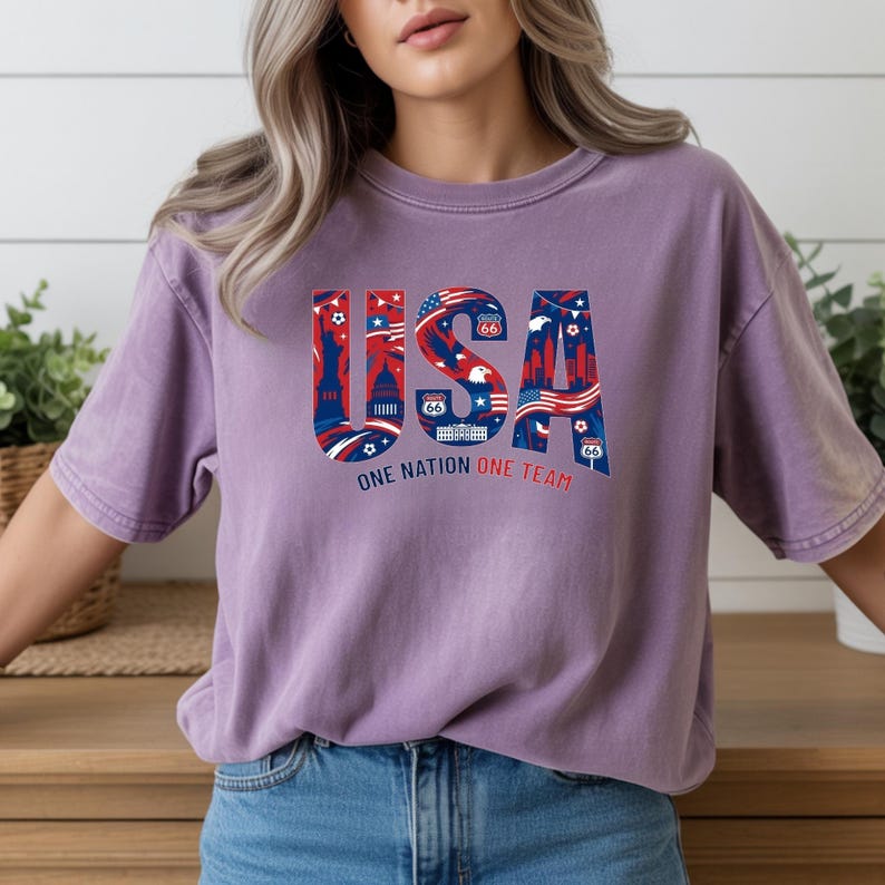 May include: A purple t-shirt with the word "USA" in red, white, and blue, featuring iconic American imagery. The text "ONE NATION ONE TEAM" is below the word "USA".