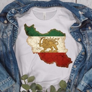 Puede incluir: Camiseta blanca con un gr&aacute;fico desgastado del mapa de Ir&aacute;n con los colores de la bandera nacional. El dise&ntilde;o incluye un le&oacute;n sosteniendo una espada, con secciones verdes y rojas. La camiseta est&aacute; debajo de una chaqueta vaquera.
