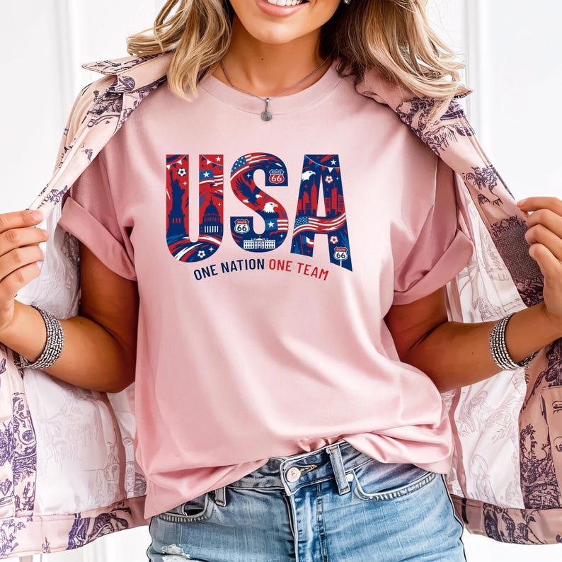 May include: A light pink t-shirt with the word "USA" in red, white, and blue, featuring iconic American imagery. The text "ONE NATION ONE TEAM" is below the word "USA". The shirt is paired with blue jeans.